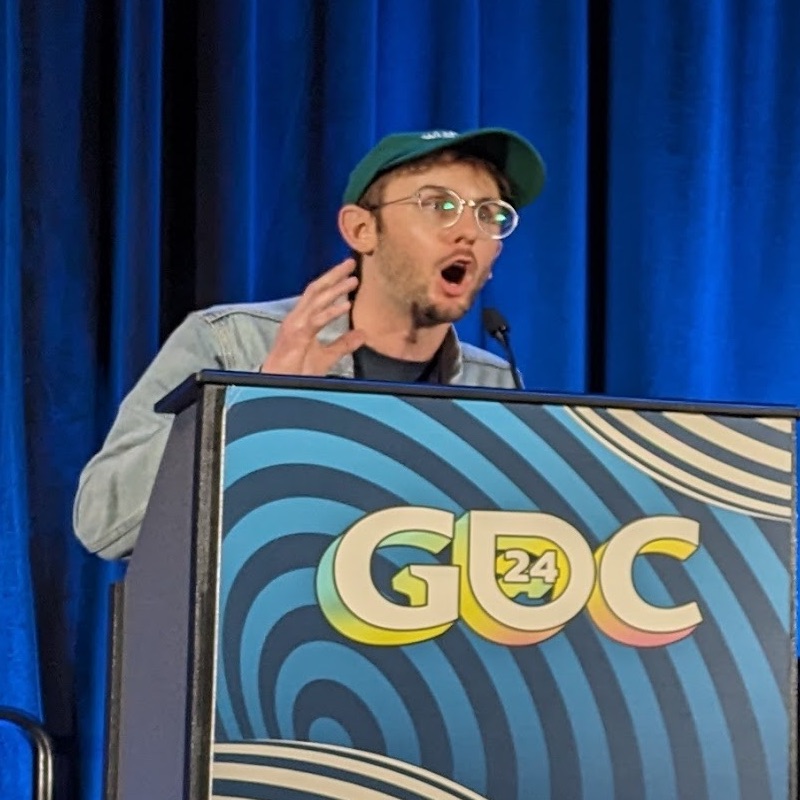 me at a podium that says 'GDC 2024.' My mouth is very open, my face is animated, and my hand is held up like I'm grabbing something.