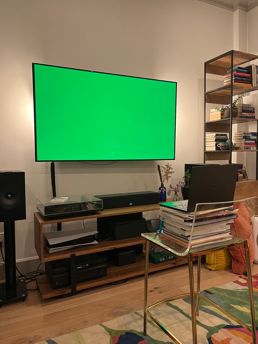 A large tv with a green image on it. A laptop sits in front of the TV on top of a chair; the TV is acting like a greenscreen.